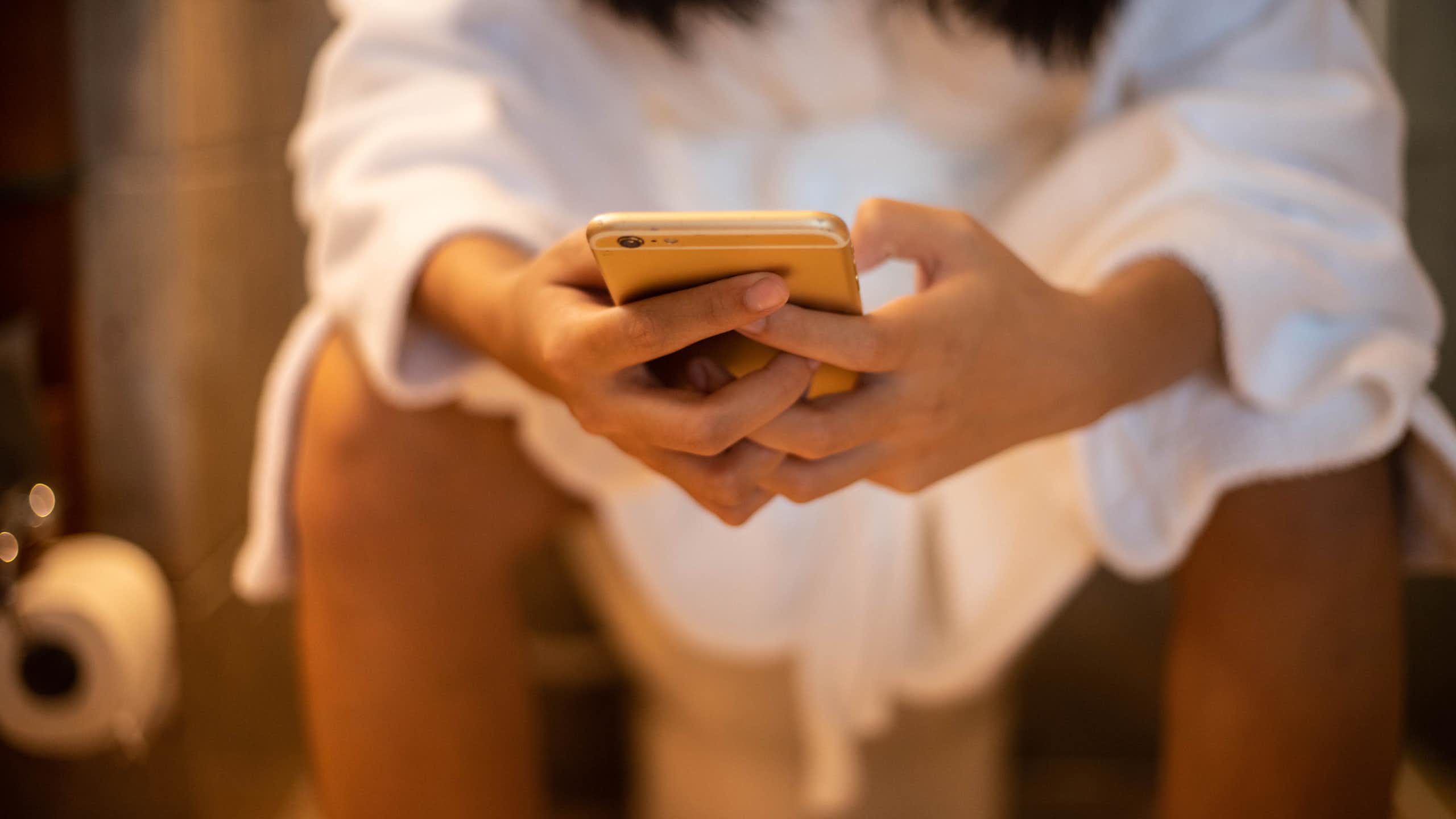 Woman sitting on toilet leaning forward hands holding smartphone