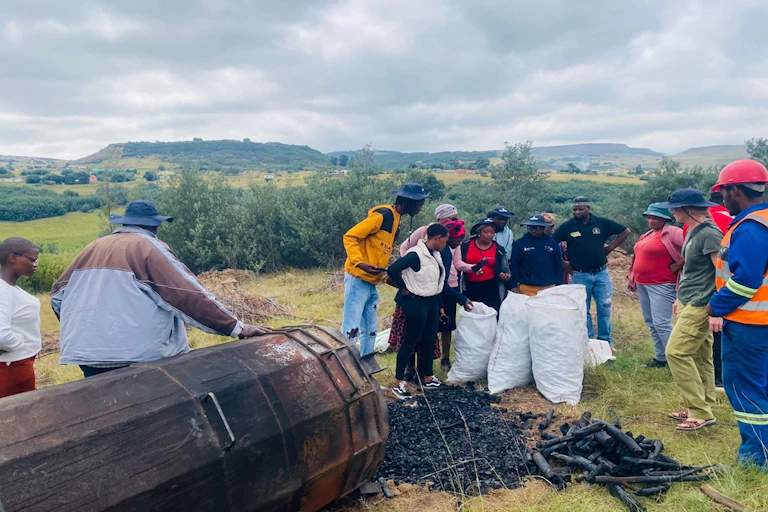 People stand around some large bags and a drum where sticks are being made into charcoal