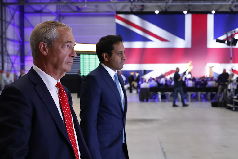 View from the side of Farage and Yusuf walking side by side, with a wall-sized union jack flag in the background