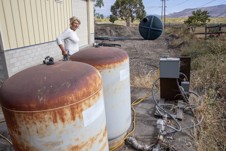A woman stands next two two water tanks with trees, mountains and field behind her.
