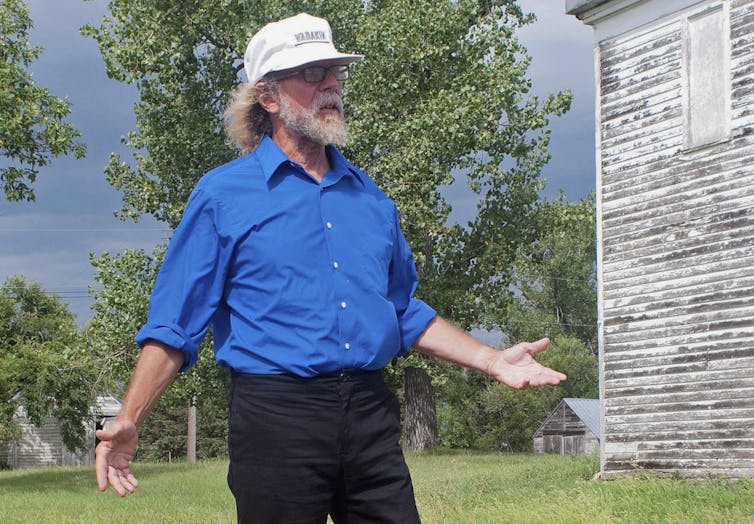 A man dressed in a blue shirt and white baseball cap stands in front of a wood house.