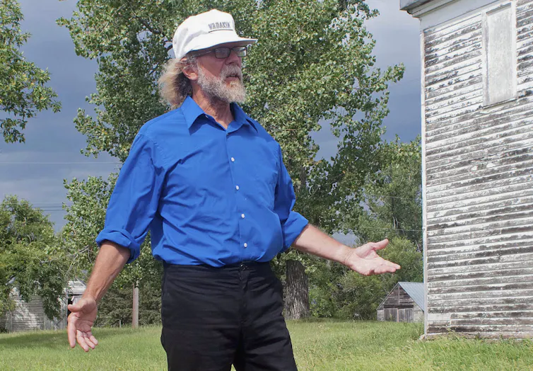 A man dressed in a blue shirt and white baseball cap stands in front of a wood house.