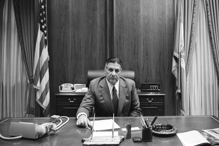 Black-and-white portrait of middle-aged man in suit sitting at large desk with American flag to his side