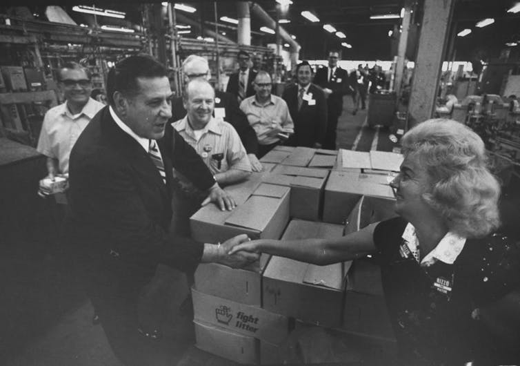 Man in suit shakes hands with woman alongside stacks of boxes while factory workers gather around