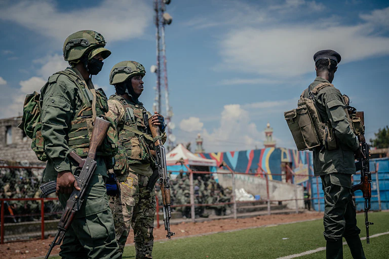 Three armed men in army fatigues standing in the foreground on a field with green turf, with a group of people seated on raised platform seats behind them