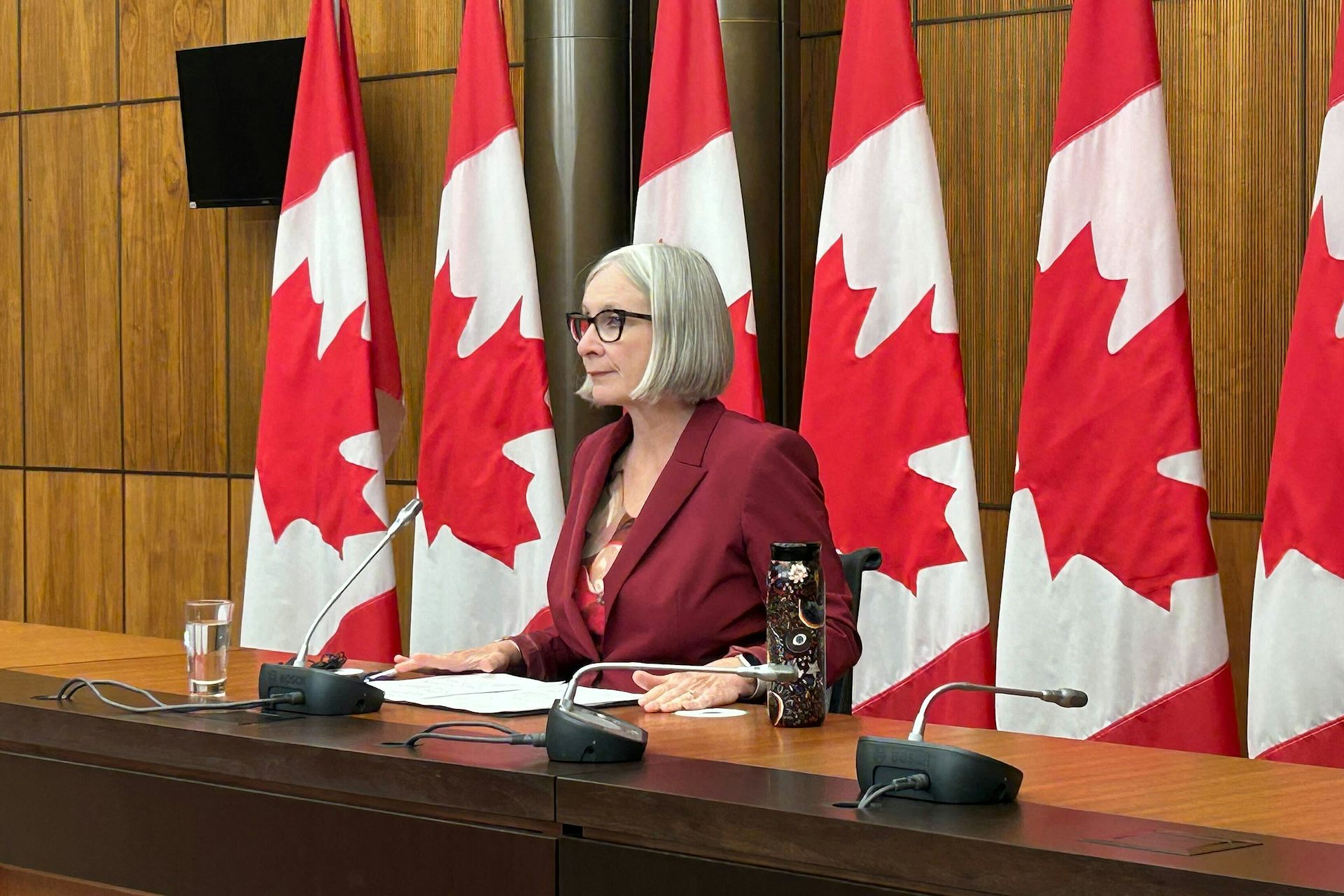 A middle-aged white woman in a red suit speaks while seated in front of a row of Canadian flags