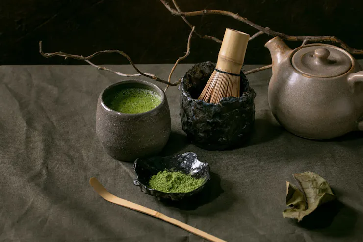 Green, frothy drink in a craft ceramic cup, next to a dish of green powder, a ceramic teapot and a bamboo whisk.