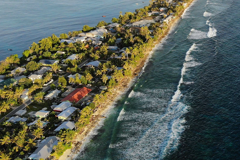 An island with homes to the edges of the water, palm trees and waves on one side.