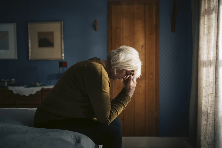 Older woman sits on a bed in dark room and holds her head in her hands.