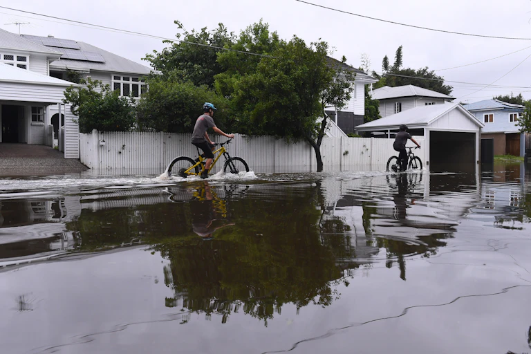 People riding bikes along a flooded street, houses in background