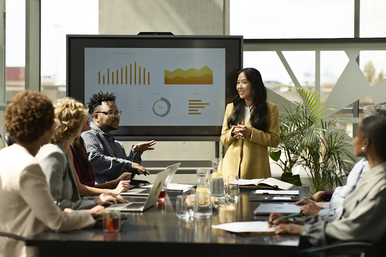 Woman standing and other people sitting around a boardroom table; a board with charts in the background