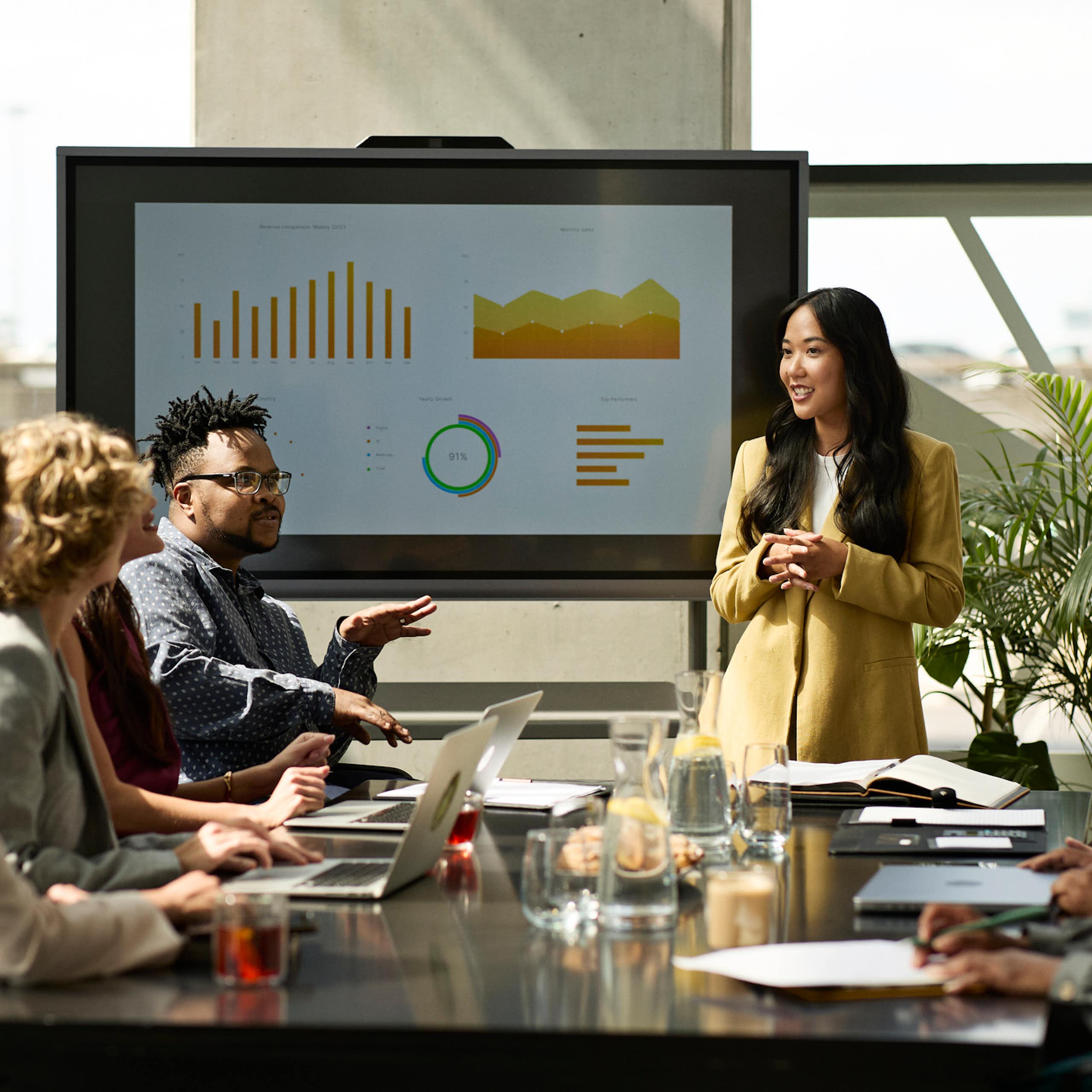 Woman standing and other people sitting around a boardroom table; a board with charts in the background