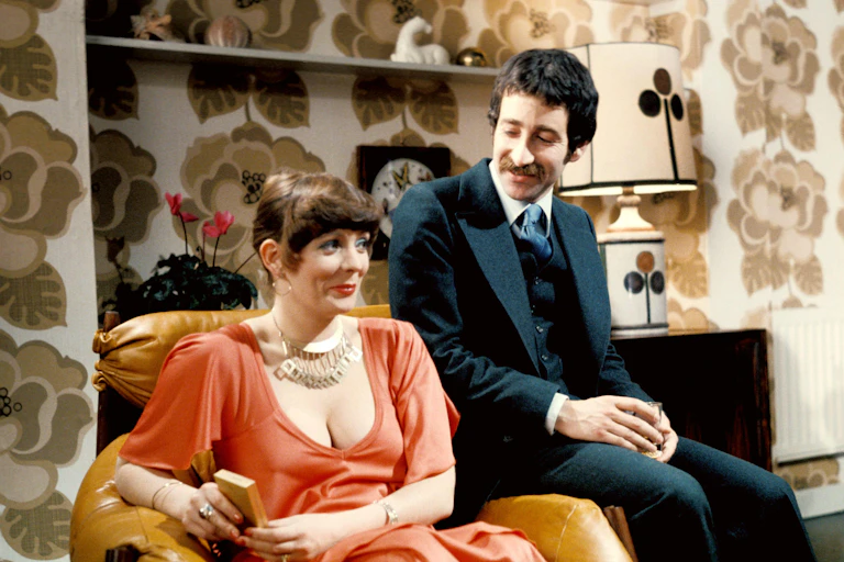 A 1970s scene at a party with a glamorous woman in an orange dress and a man perched on the arm of her chair looking down at her.