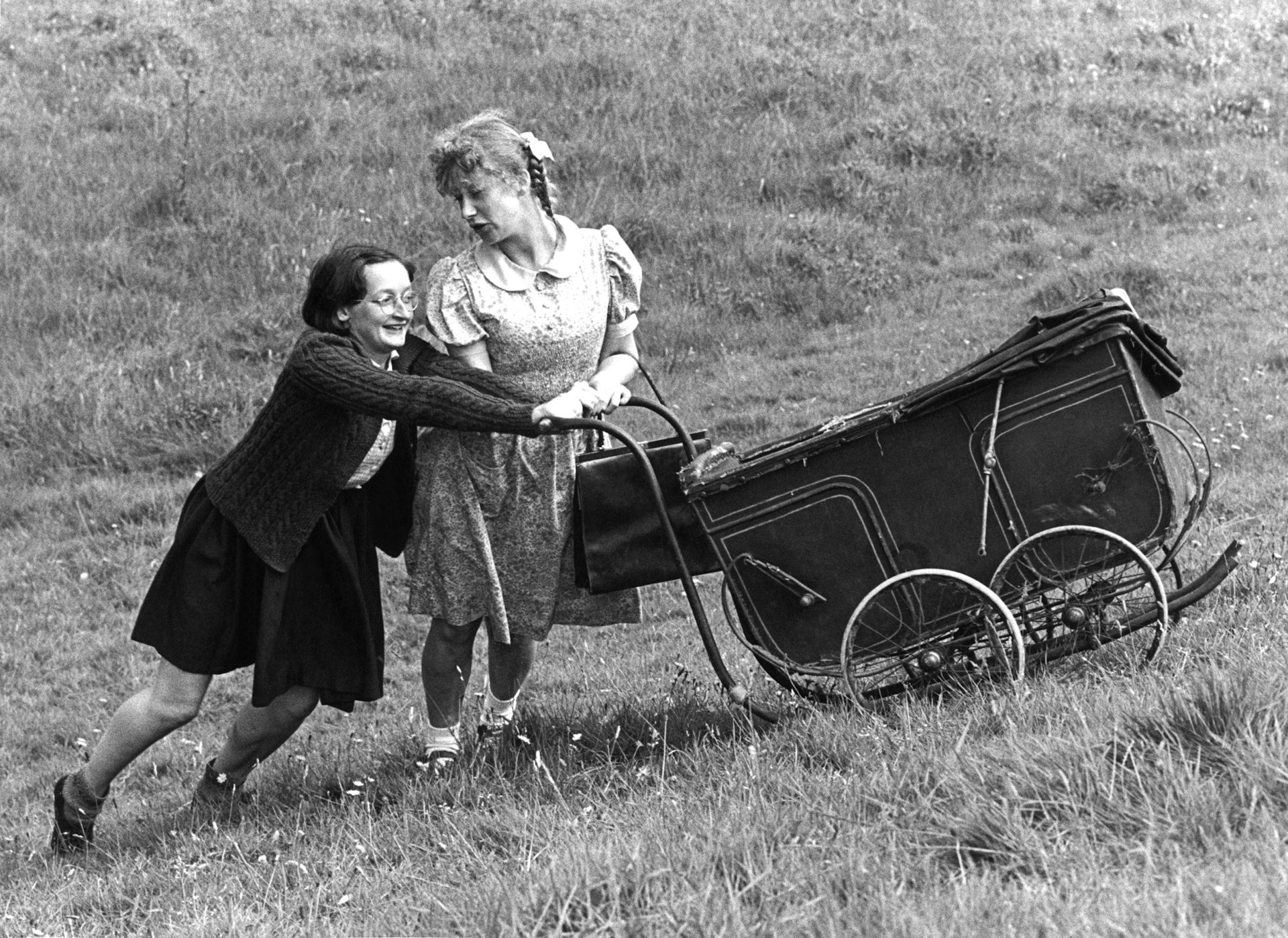 Two women acting as children, pushing a 1940s style pram up a hill.
