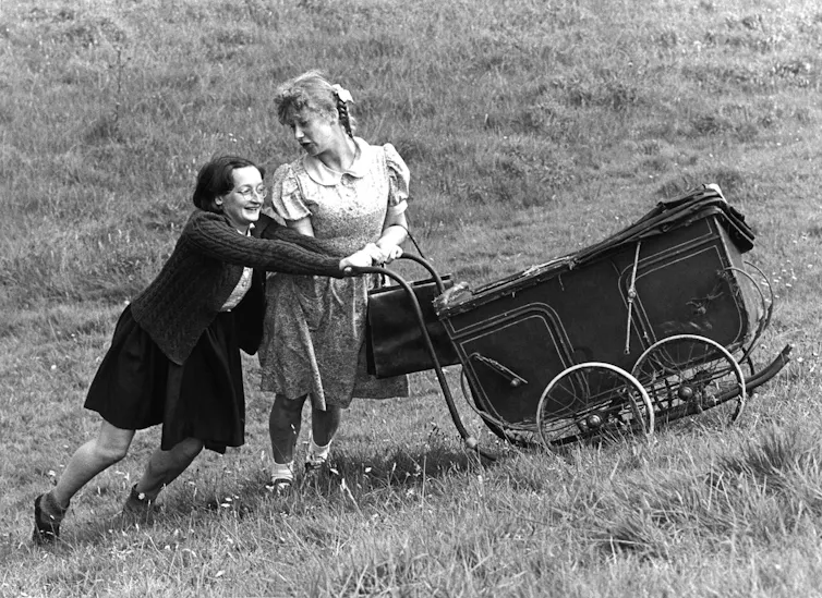 Two women acting as children, pushing a 1940s style pram up a hill.