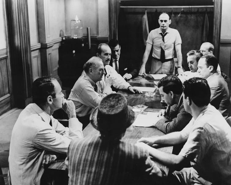Black-and-white photo of ten men sitting around a table and one standing at the head, addressing the group.