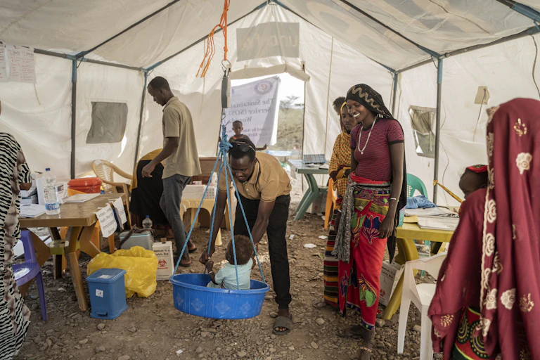 Women look on as a man weighs a baby inside a tent