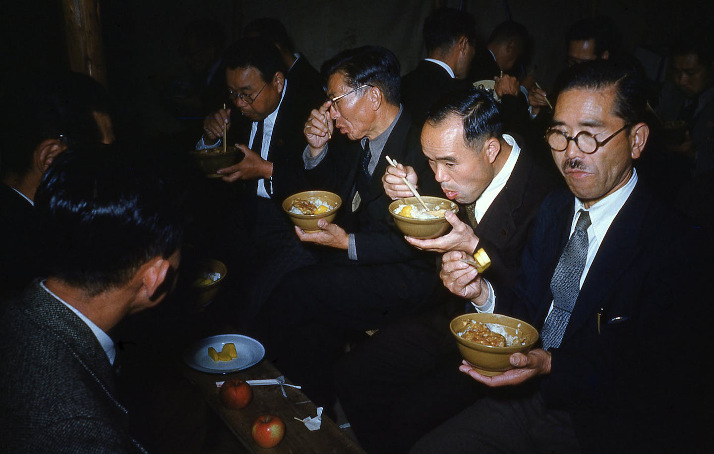 Japanese men eating ramen, rice, and eggs with chopsticks, dressed in formal suits, wearing glasses, with apples and other food on a table, in a crowded room,