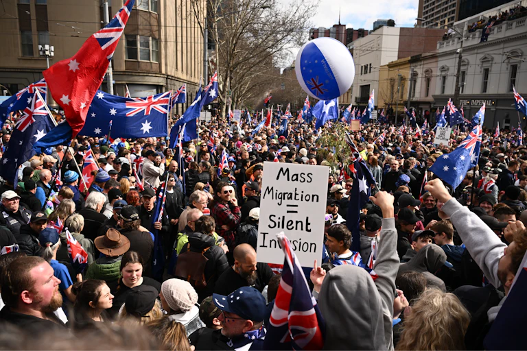 A large protest crowd waving Australian flags and a sign that says mass migration equals silent invasion