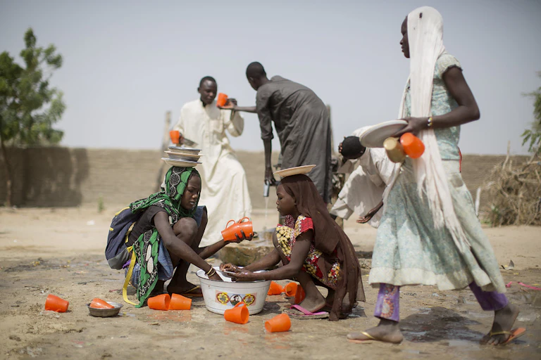 Girls squatting on the sandy ground in front of a basin, with numerous plastic mugs, and adults carrying mugs nearby