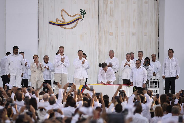 Hombres y mujeres vestidos de blanco en un escenario firman un acuerdo ante gente que les observa desde el público.