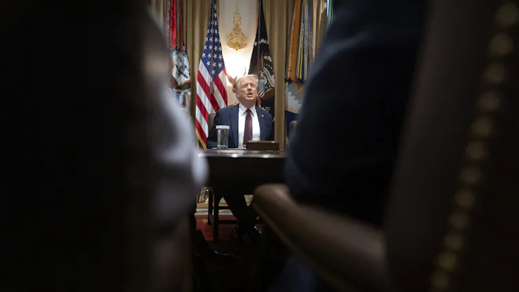 A man sits at an ornate desk photographed between the arms of people standing in front of him.