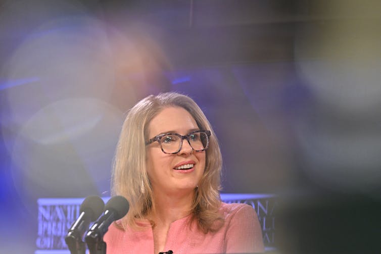 A blonde woman in glasses smiles in front of a blue background