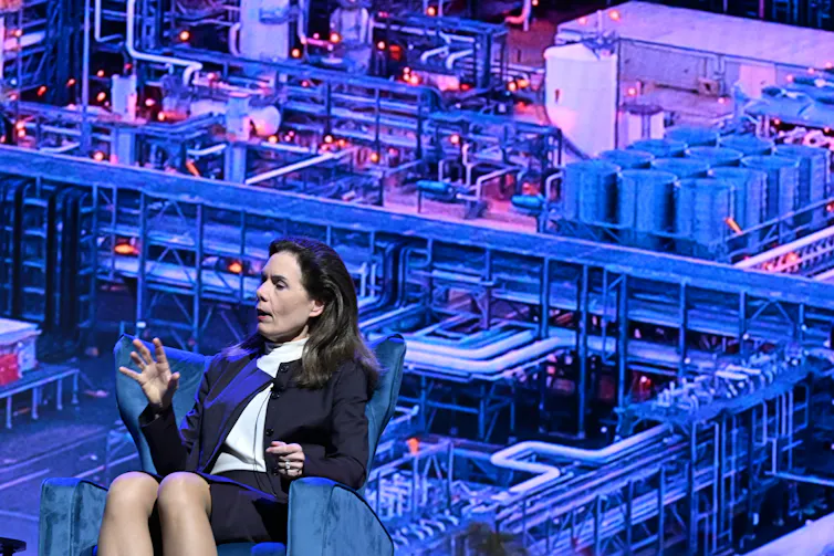 a woman sits against a background of pipes and tanks