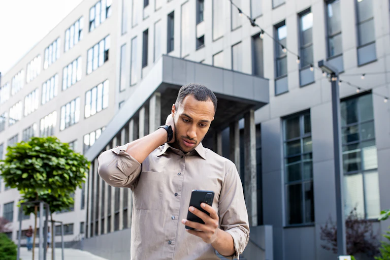 Stressed man rubbing head with hand while looking at mobile phone screen on background of office building.