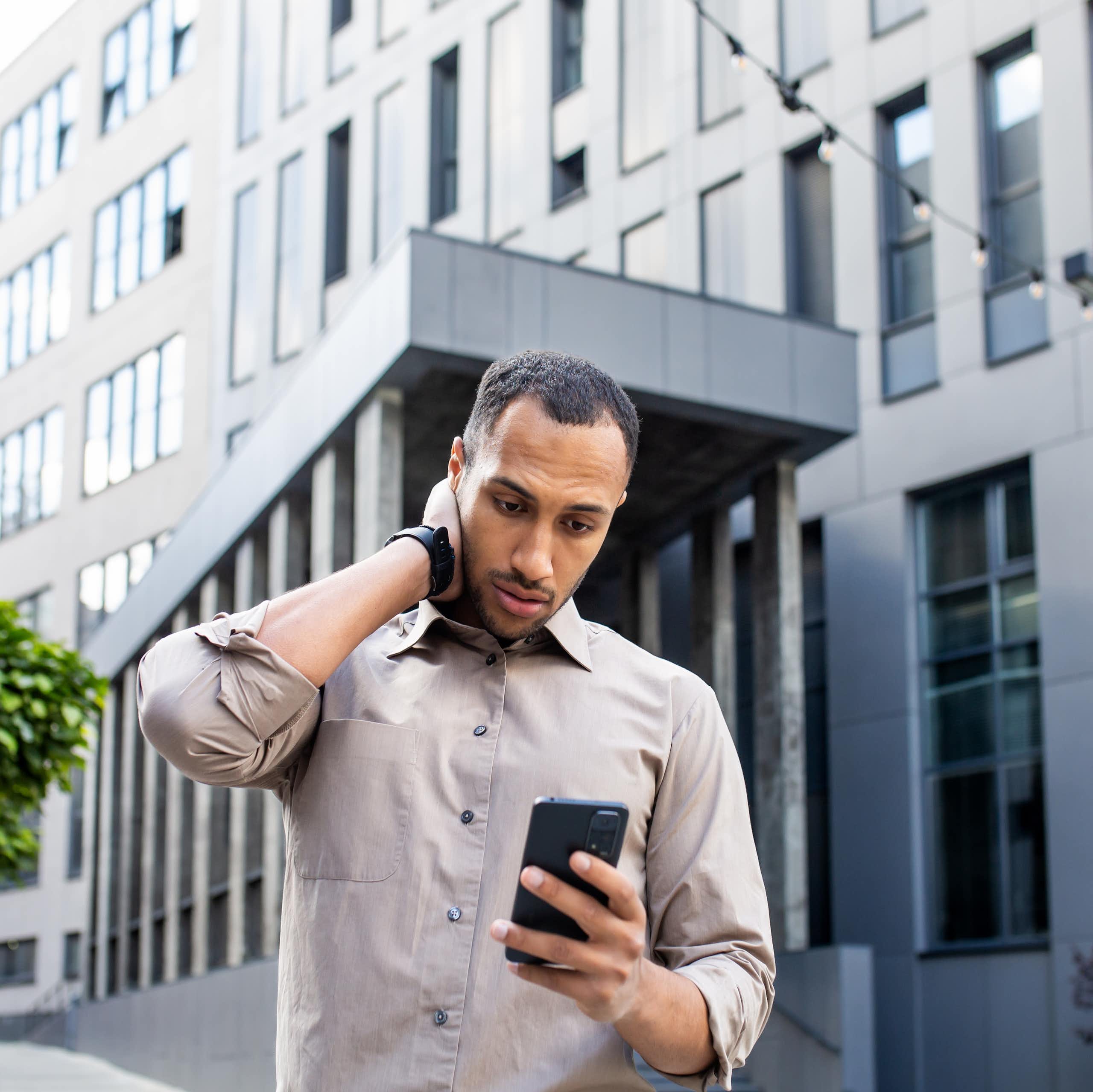 Stressed man rubbing head with hand while looking at mobile phone screen on background of office building.