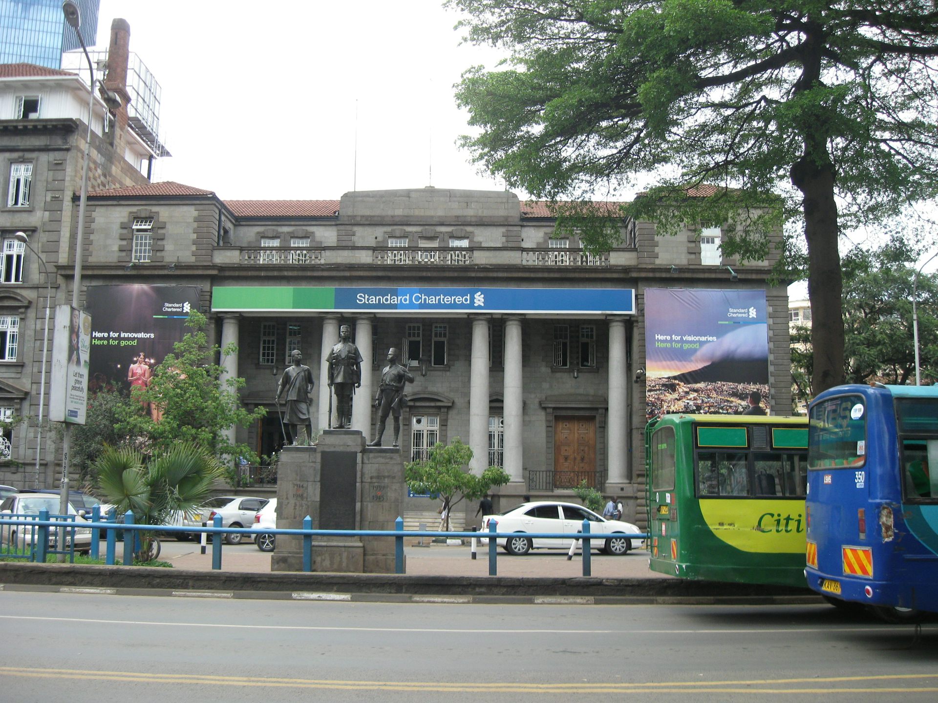 The front of a bank building foregrounded by a monument and street traffic