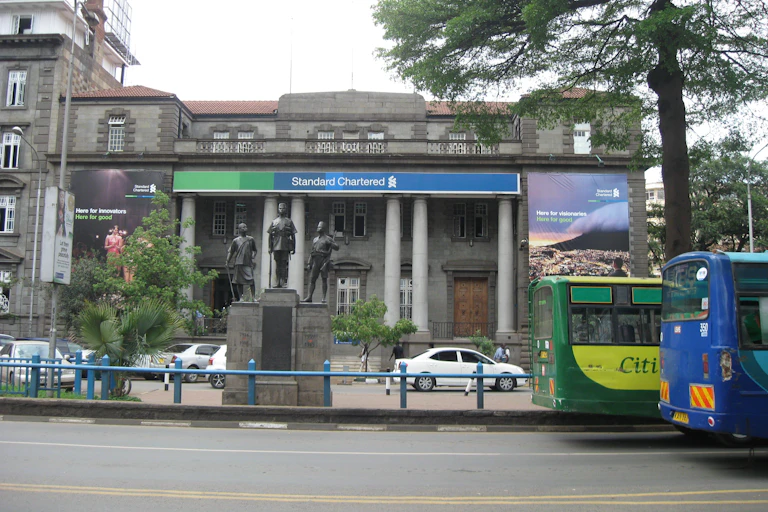 The front of a bank building foregrounded by a monument and street traffic