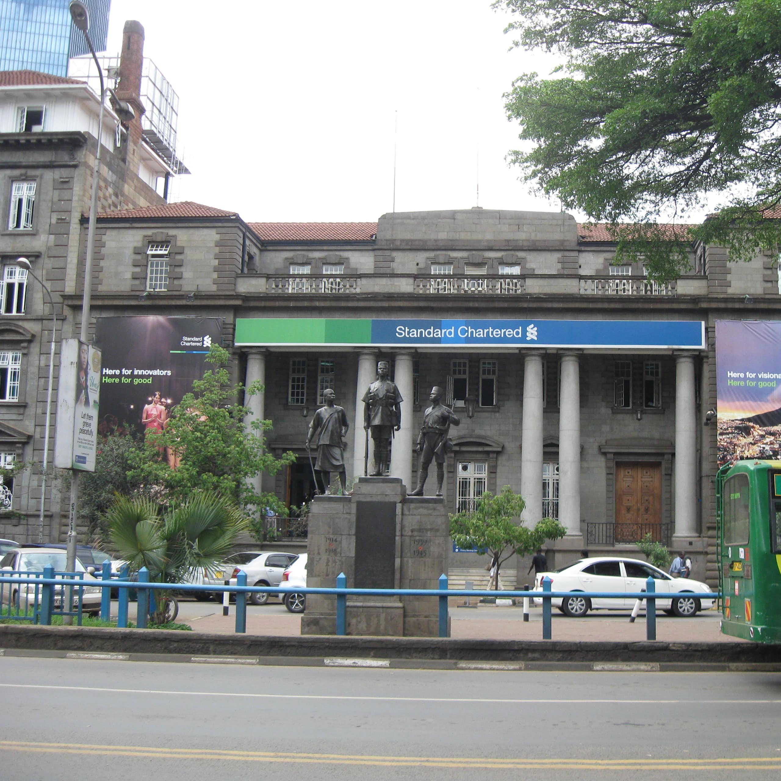 The front of a bank building foregrounded by a monument and street traffic