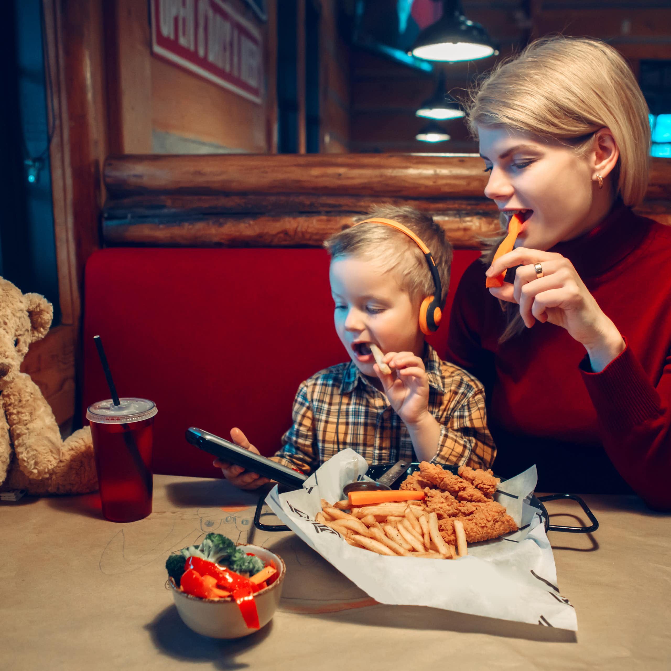 Mujer y niño comiendo en un restaurante mientras miran la pantalla de un dispositivo.