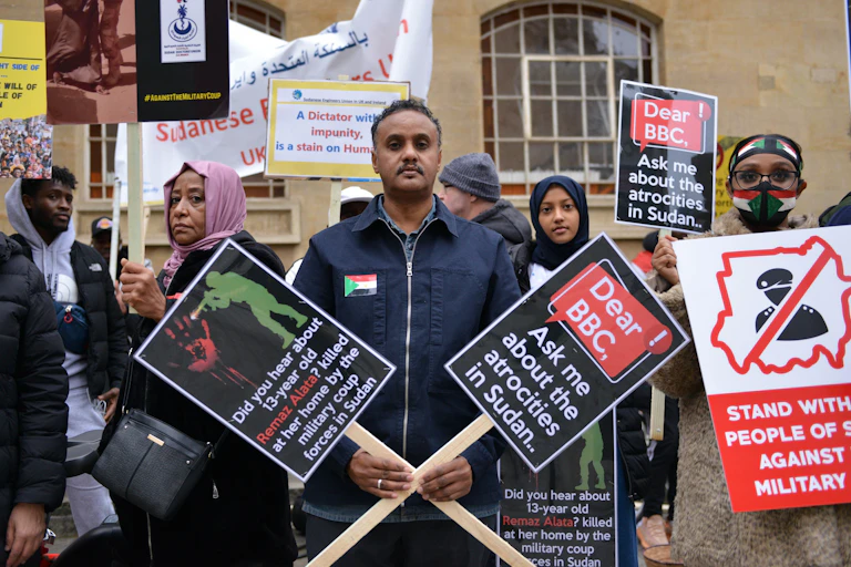 A man holds two posters, crossed in front of him, one reading "Dear BBC, ask me about the atrocities in Sudan". There are other people with placards behind him.