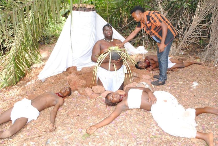 A man directs actors dressed in simple white outfits. One kneels holding a sacred object, others lie passed out on the ground.
