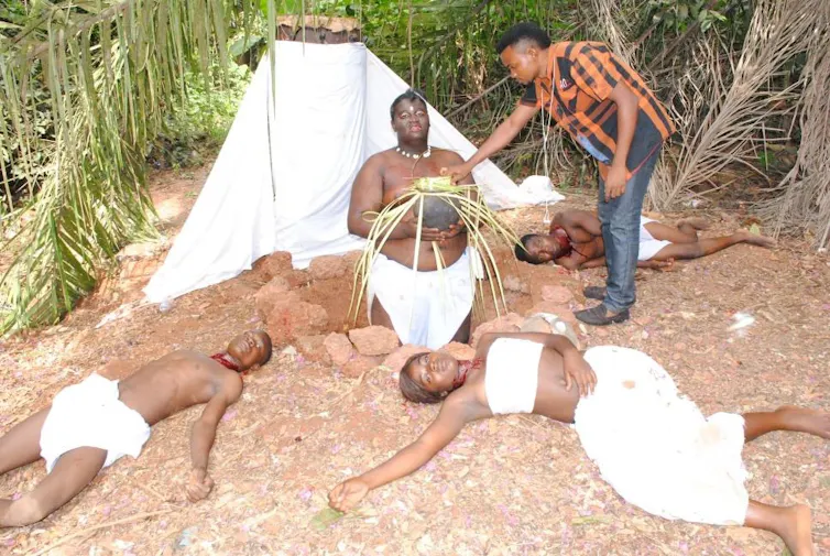 A man directs actors dressed in simple white outfits. One kneels holding a sacred object, others lie passed out on the ground.