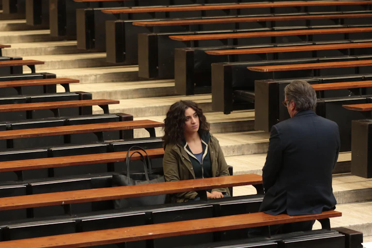 Una alumna sentada habla con un profesor en un aula vacía.