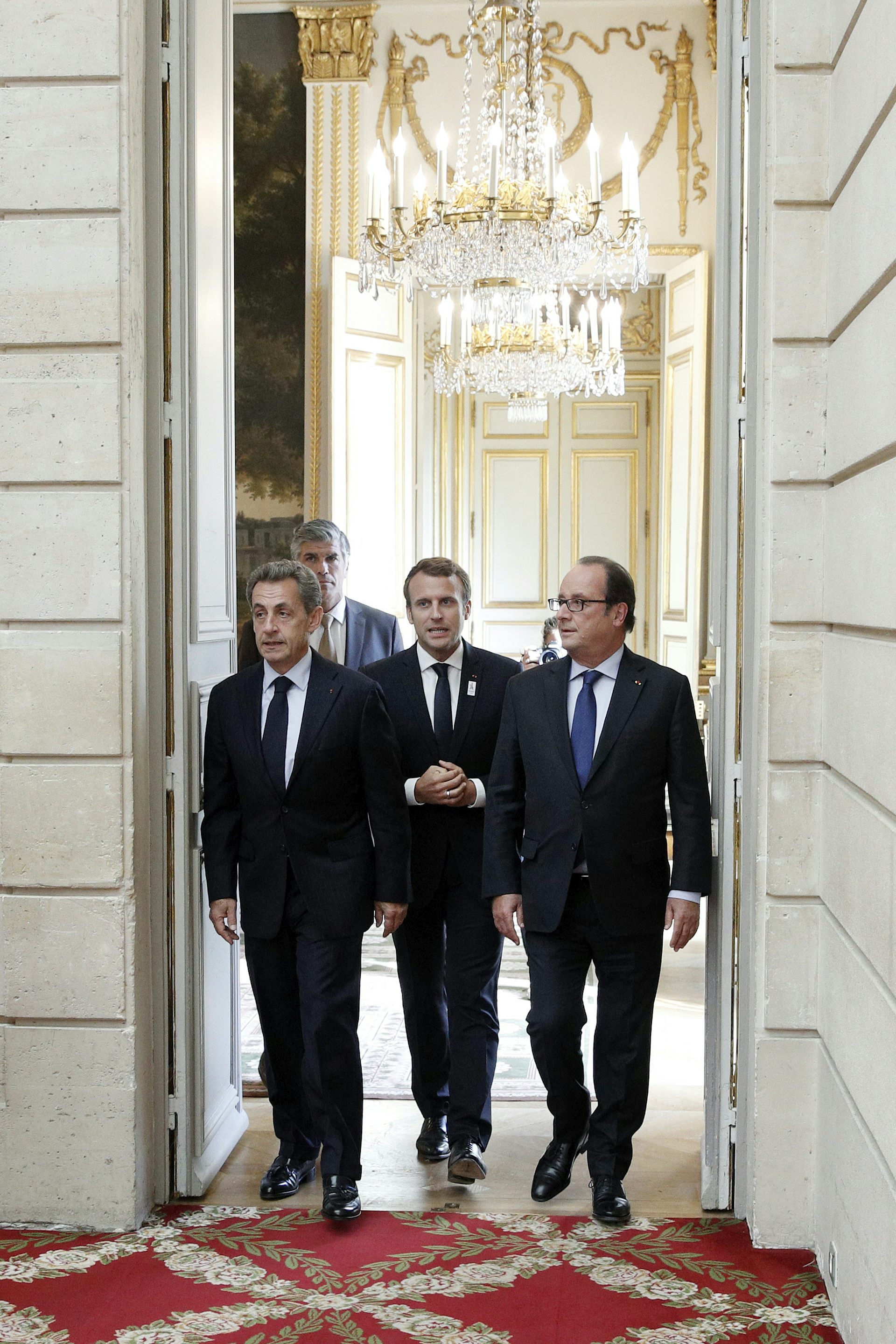 From left to right, former French president Nicolas Sarkozy, current President Emmanuel Macron, and former president François Hollande appear in front of an open door.