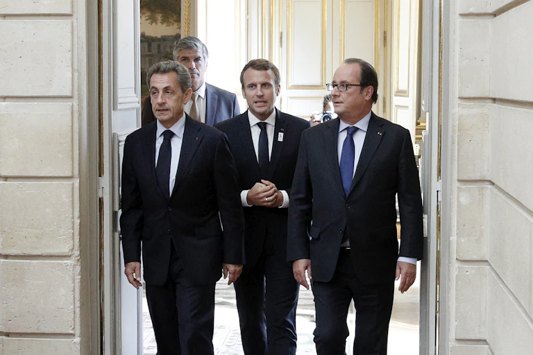 From left to right, former French president Nicolas Sarkozy, current President Emmanuel Macron, and former president François Hollande appear in front of an open door.