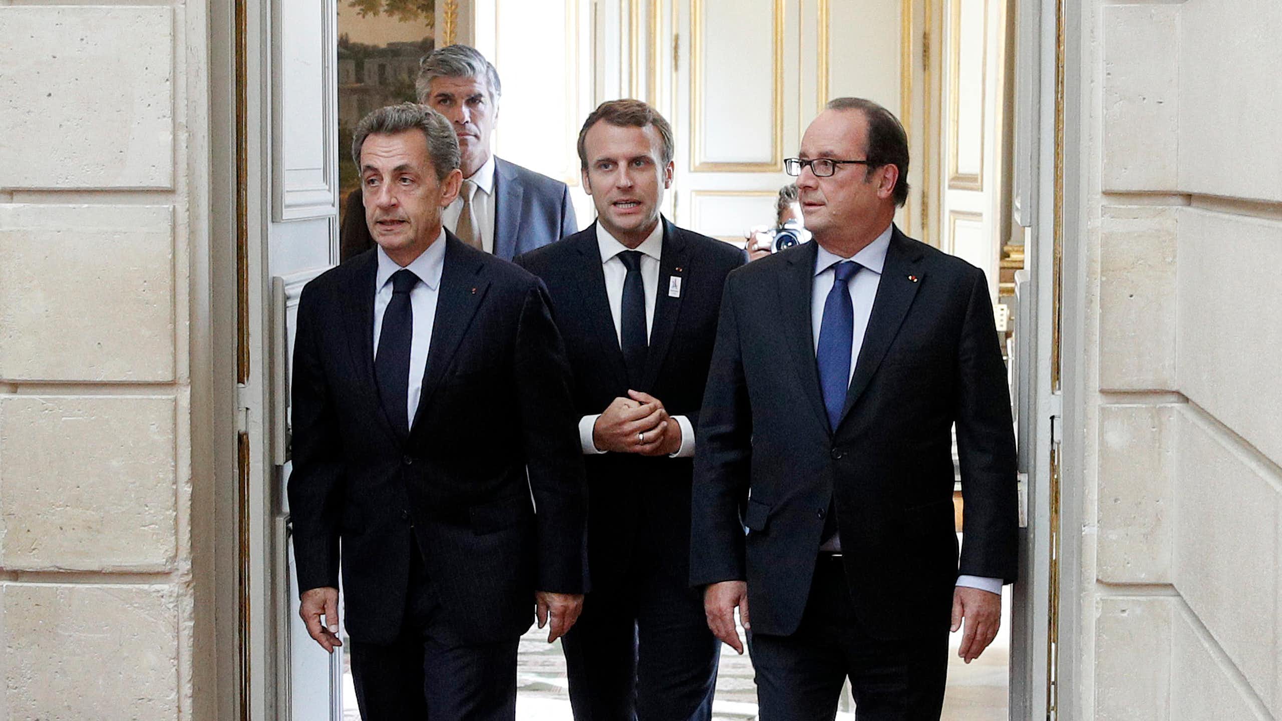 From left to right, former French president Nicolas Sarkozy, current President Emmanuel Macron, and former president François Hollande appear in front of an open door.