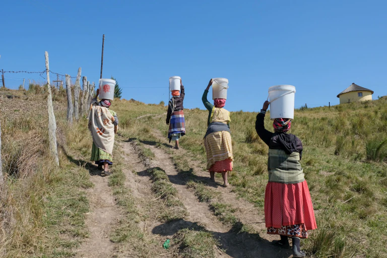 Four elderly women walk up a steep and grassy hill on a dirt track, each with a 20 litre bucket of water balanced on their heads