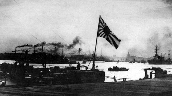 A flag is seen above a smoking boat and buildings.