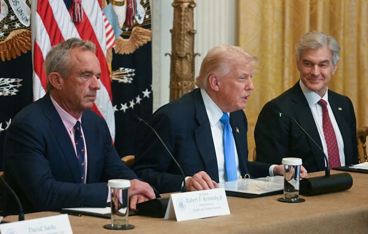 RFK Jr, Donald Trump and Dr. Oz seated at a table with flags behind them