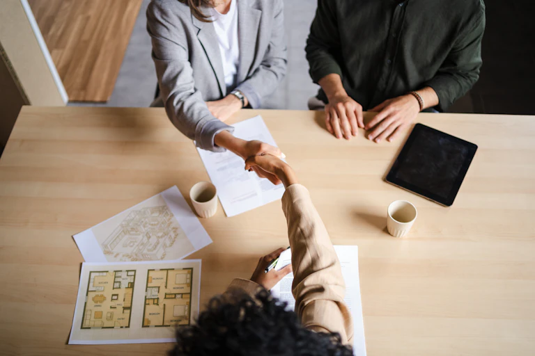 Two people in businesswear are photographed from above shaking hands. They are sitting at a table covered with architectural plans, documents and cups of coffee.