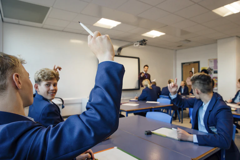 Students raise their hands in class. A teacher stands by a whiteboard in the background.