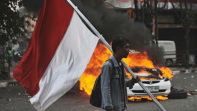 一名男子手持印尼國旗,背景中一輛汽車正在燃燒