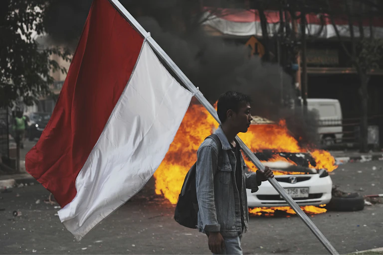 Man holds an Indonesian flag while a car burns in the background