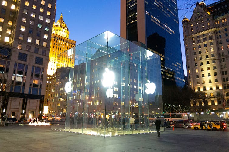 A large glass cube lit up at night with glowing Apple logos on the sides and stairs leading down to the store below.