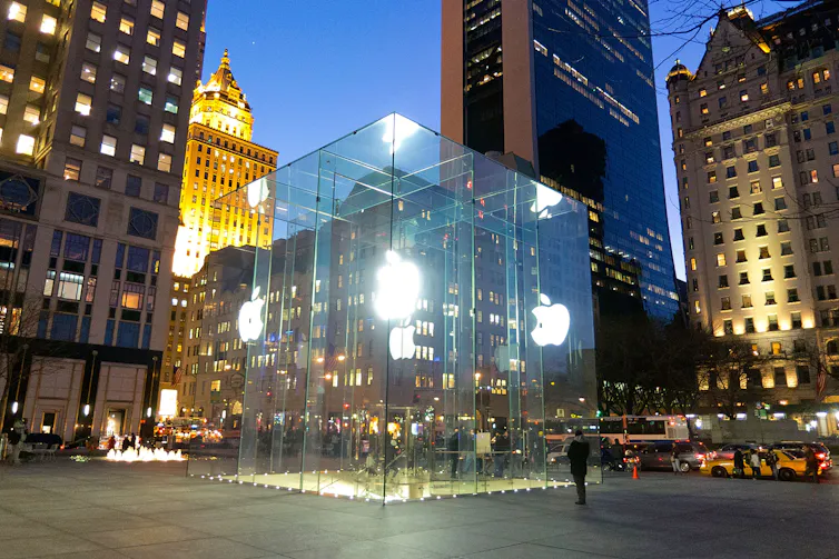 A large glass cube lit up at night with glowing Apple logos on the sides and stairs leading down to the store below.